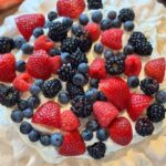 Classic Pavlova topped with whipped cream, strawberries, blueberries, raspberries, and blackberries on a wooden serving board.