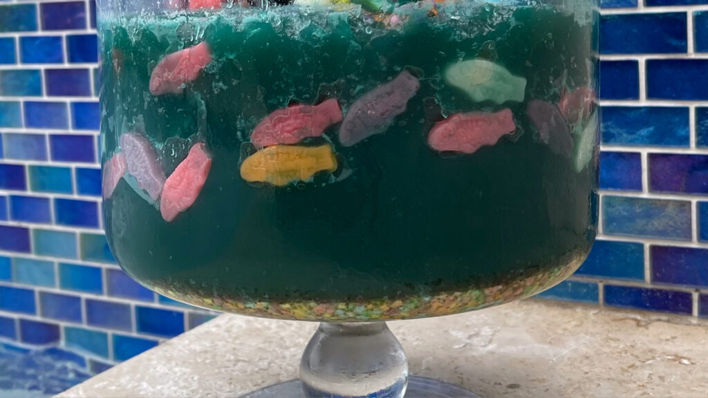 Blue gelatin dessert in a glass bowl with colorful gummy fish and candy sprinkles resembling aquarium gravel.