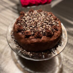 A gluten-free chocolate layer cake frosted with rich chocolate buttercream, decorated with piped chocolate roses around the edge, and topped with a generous layer of chocolate curls on a glass cake stand.