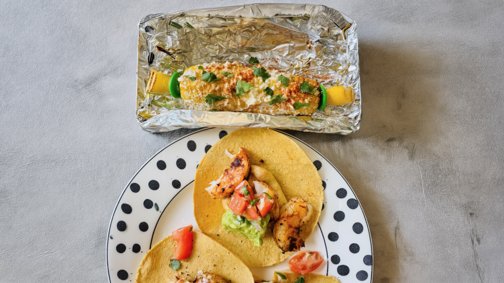 Mexican elote topped with creamy sauce, cheese, chili seasoning, and cilantro in a foil tray beside shrimp tacos.