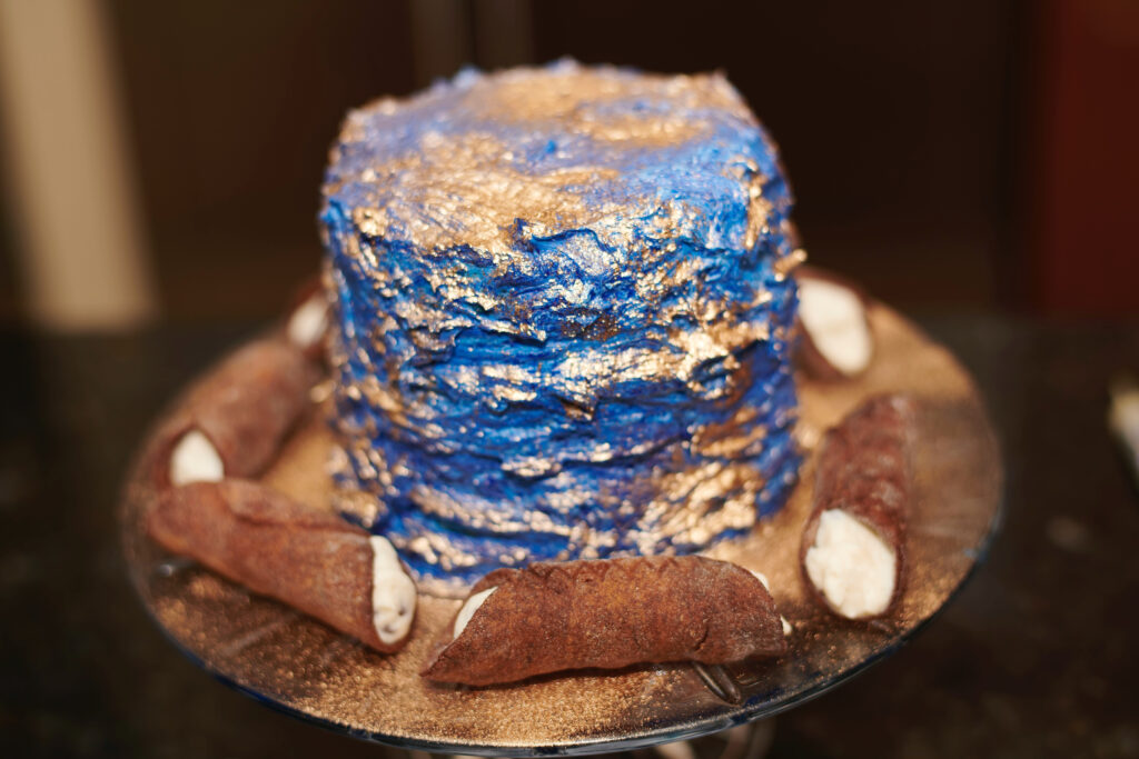 Gluten-free chocolate cannoli filled with sweet cream and arranged around a blue and gold decorated cake.