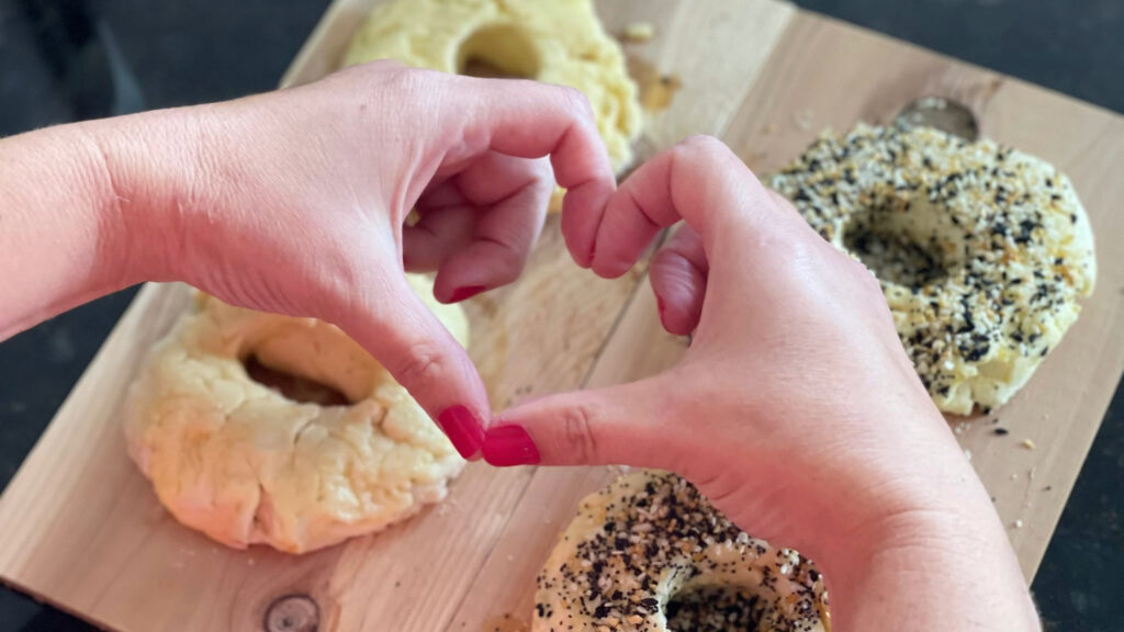 Hands forming a heart above homemade plain and everything-seasoned bagels on a wooden board.