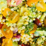 A colorful bowl of fresh avocado corn salad made with sweet corn, diced avocado, cherry tomatoes, cucumber, red onion, fresh cilantro, parsley, and homemade lime dressing, garnished with cilantro.