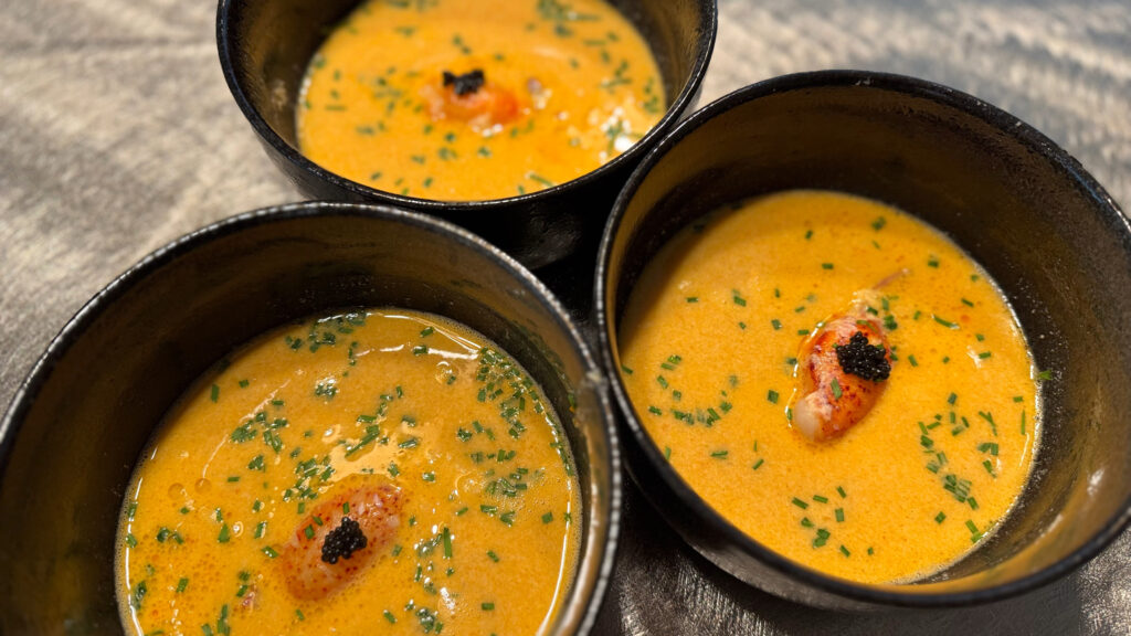 Three black bowls of creamy lobster bisque garnished with lobster meat, chopped chives, and black caviar.
