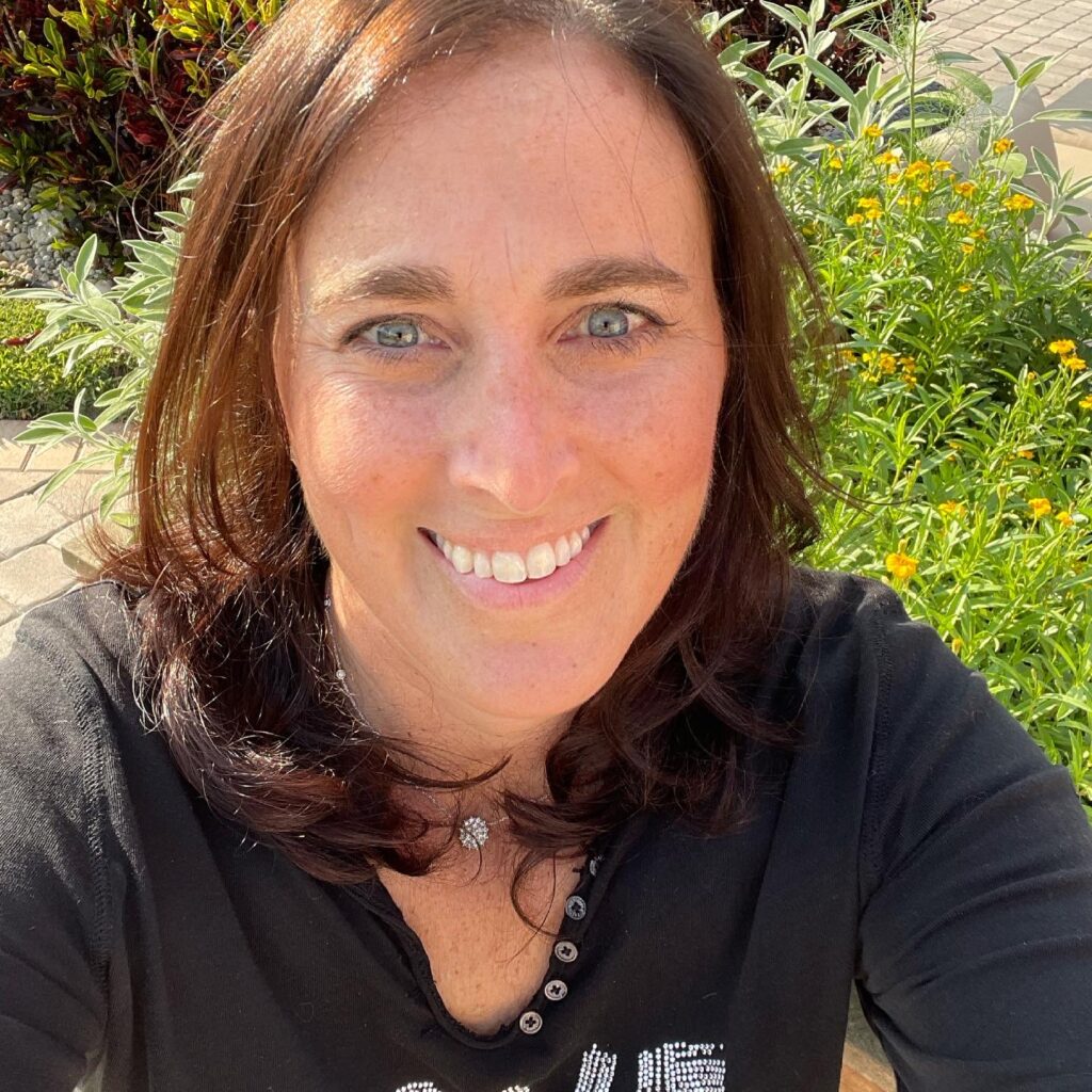 Leslie, founder and creator of Culibloom, smiling in her sunlit garden among green foliage and yellow flowers.